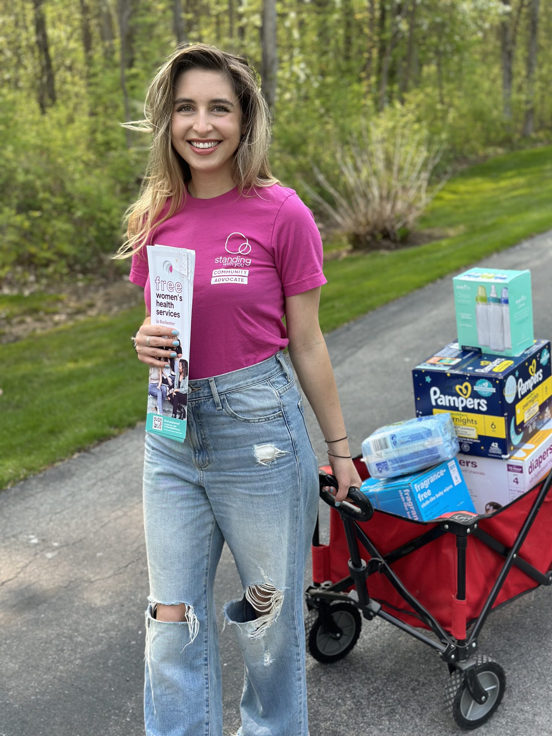 A woman in a red shirt smiles while pulling a wagon that contains baby supplies.
